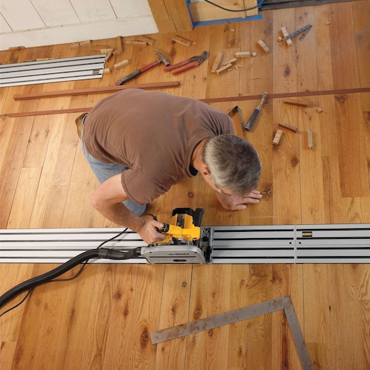A person using a DEWALT track saw, model DWS520K, to cut wooden flooring. The saw is positioned on a metal track, and there are woodworking tools and wood scraps scattered on the floor.