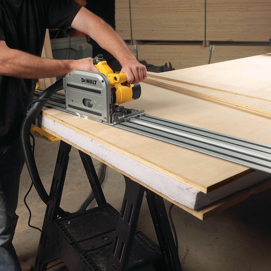 A person using a DEWALT track saw to cut a large sheet of wood, with the saw guided along a metal rail placed on the work surface.
