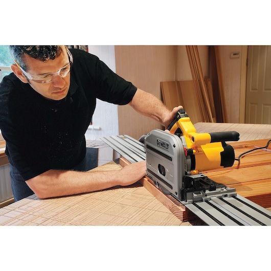A person using a DEWALT DWS520K track saw to cut a wooden board in a workshop. The saw is positioned on a metal guide rail for precise cutting. The person's face is blurred for privacy.