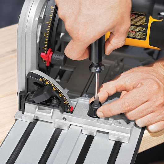 Close-up of a person using a screwdriver to adjust a DEWALT track saw, with visible measurement markings and adjustment knobs. The saw is set on a wooden surface.