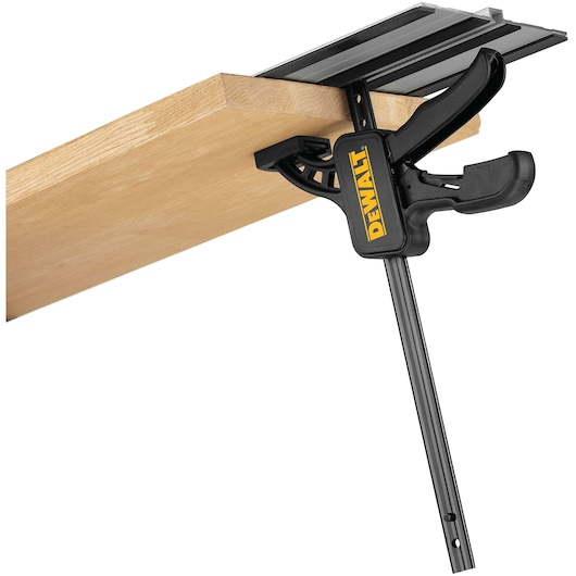 A DEWALT branded clamp is attached to the edge of a wooden board, holding a black guide rail in place. The clamp is black and features the DEWALT logo in yellow.