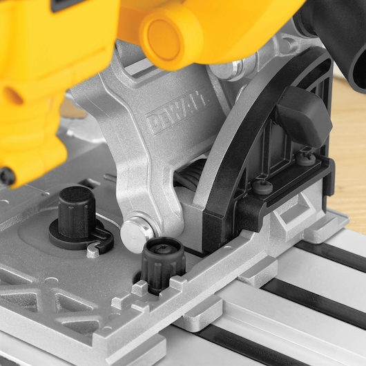 Close-up view of a DEWALT DWS520K track saw, highlighting its metal base, adjustment knobs, and part of the handle. The saw is shown resting on a grooved surface, commonly used for precision cutting.