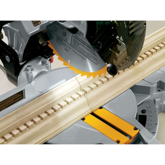 Close-up of a DEWALT miter saw cutting a piece of decorative wood trim. The saw blade is making a precise angled cut, and the wood trim has a detailed geometric pattern.