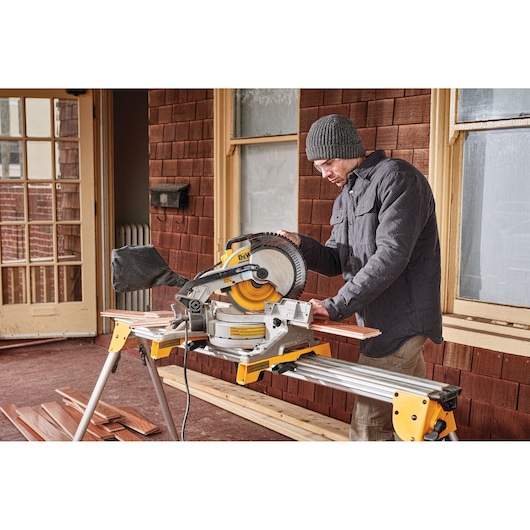 A person using a DEWALT DWS713 miter saw to cut wooden boards on a stand, wearing a jacket and beanie, in front of a building with brown shingles and windows.