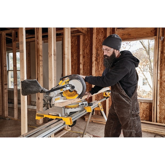 A person wearing work overalls and a hat is operating a DEWALT DWS713 miter saw to cut a piece of wood inside a partially constructed building.
