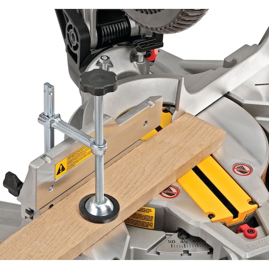 A close-up view of a DEWALT miter saw (model DWS713), showing the saw securely clamping a piece of wood for precise cutting. Visible elements include the saw blade, metal clamp, yellow safety warnings, and adjustment dials.