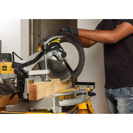 A person wearing gloves is using a DEWALT DWS715 miter saw to cut a wooden plank in an indoor workshop. The saw is mounted on a yellow workbench and surrounded by other tools and equipment.