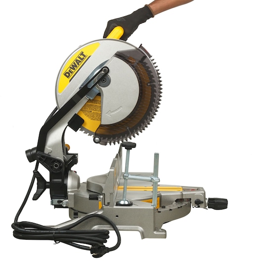 A DEWALT DWS715 miter saw with a yellow and silver body, shown in use by a gloved hand. The saw is plugged in and positioned for cutting, with visible blade, handle, and base.