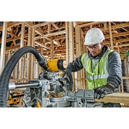 A worker wearing a safety vest and hard hat operates a DEWALT DWS727 miter saw in a wooden frame construction site. The saw is connected to a dust extraction hose.