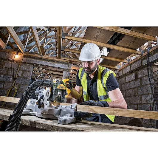 A person wearing a safety helmet and high-visibility vest is operating a DEWALT DWS774 miter saw to cut a wooden plank inside a construction site with exposed beams and brick walls.