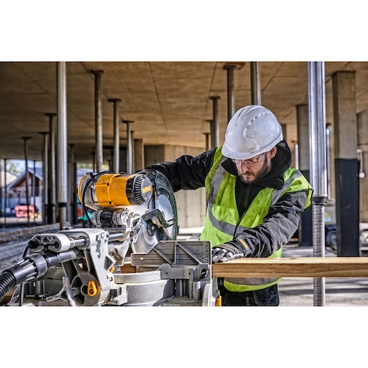 A construction worker wearing a safety vest and hard hat is using a DEWALT DWS780 miter saw to cut a piece of wood at an outdoor construction site.