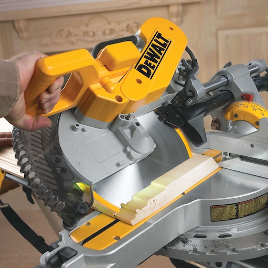 A close-up of a person operating a DEWALT DWS780 miter saw, cutting a piece of wood with the circular blade in a woodworking environment.