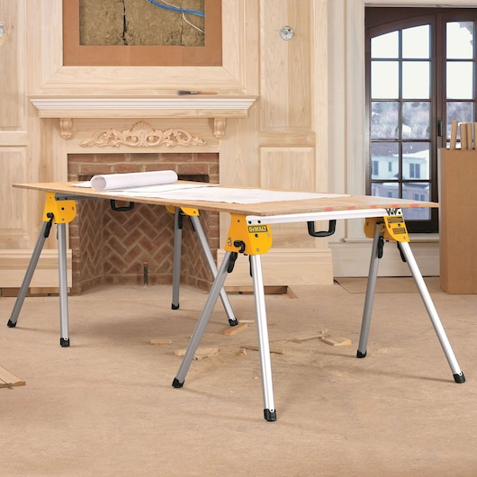 A DEWALT DWS780 yellow and silver sawhorse set used as a work table, with a wooden board and rolled plans on top, placed in a room under renovation with unfinished woodwork and a fireplace.