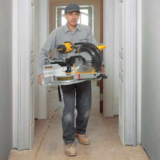 A person carrying a DEWALT DWS780 miter saw in a hallway during construction or renovation. The individual is wearing work clothes and boots, with the face blurred for privacy.