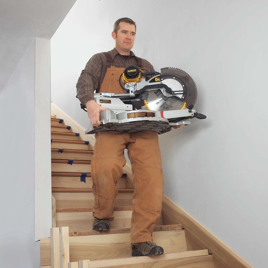 A person in brown work overalls is carrying a DEWALT DWS780 miter saw up a staircase inside a building. The face of the person is blurred for privacy.