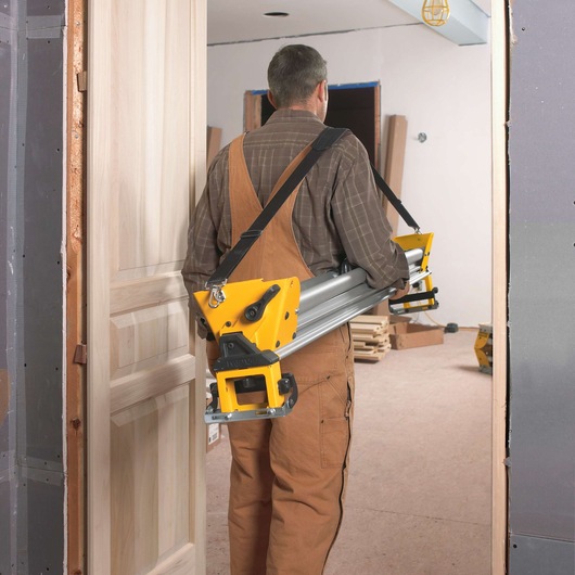 A person wearing brown overalls and a plaid shirt is carrying a DEWALT miter saw stand through a doorway in a room under construction.