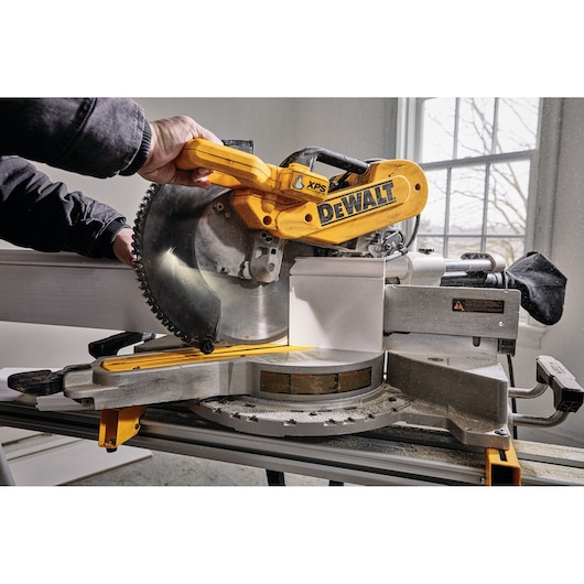 A close-up view of a DEWALT DWS780 miter saw being used by a person to cut a piece of wood indoors. The saw is mounted on a workbench, with natural light coming in from a window in the background.