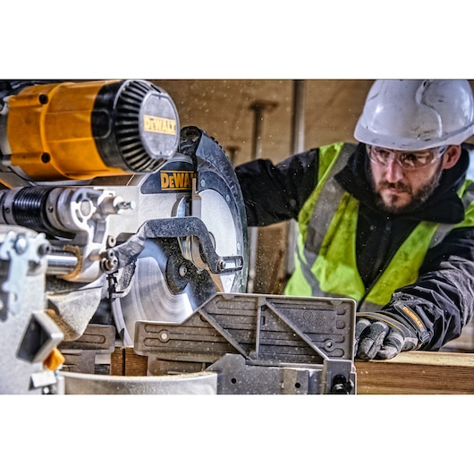 A DEWALT DWS780 miter saw being used by a person wearing a safety vest, gloves, and a hard hat. The saw is cutting through a piece of wood at a construction site.