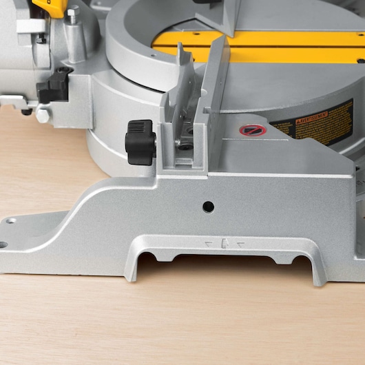 Close-up view of a DEWALT DWS780 miter saw, showing the metallic base, adjustment knob, and part of the yellow cutting guide on a light wooden surface.