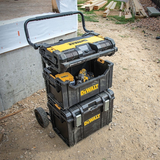 A stackable DEWALT mobile tool storage system with three black and yellow toolboxes on rugged wheels and a handle, shown outdoors at a construction site. The middle box contains various tools, including a cordless drill.