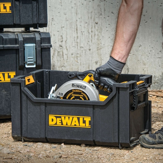 A person wearing gloves places a DEWALT circular saw inside a black DEWALT tool storage box outdoors. Other DEWALT storage boxes are visible in the background.