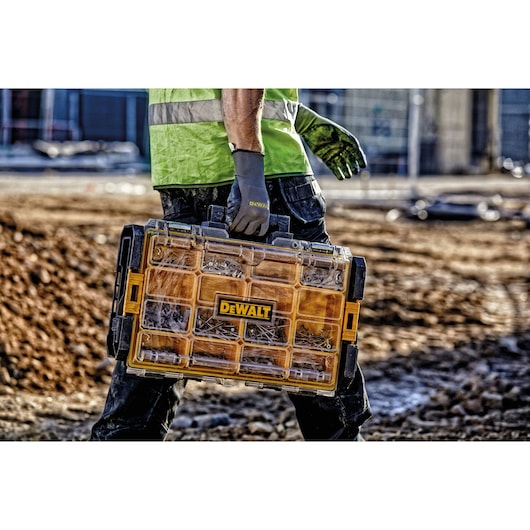 A person wearing work gloves and a high-visibility vest is carrying a DEWALT organizer case filled with various screws and fasteners at a construction site.