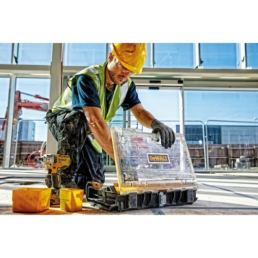 A person wearing a yellow hard hat, high-visibility vest, and gloves is on a construction site, opening a transparent DEWALT storage box. A DEWALT power tool and yellow containers are nearby on the floor.