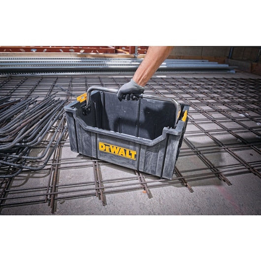 A person wearing a glove is lifting a black DEWALT toolbox with a metal handle. The toolbox is placed on a construction site floor with metal rebar grids.