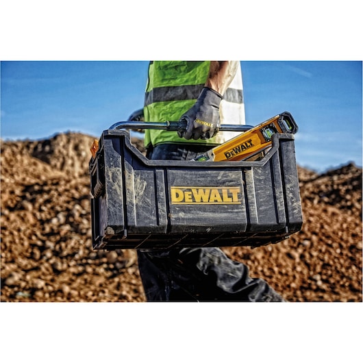 A person wearing work gloves and a high-visibility vest carries a DEWALT toolbox containing tools, including a yellow DEWALT spirit level, at an outdoor construction site with dirt piles in the background.