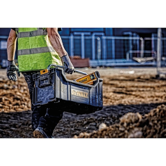 A person wearing a high-visibility vest and gloves carries a DEWALT tool box with tools, including a yellow spirit level, on a construction site with dirt ground and fencing in the background.