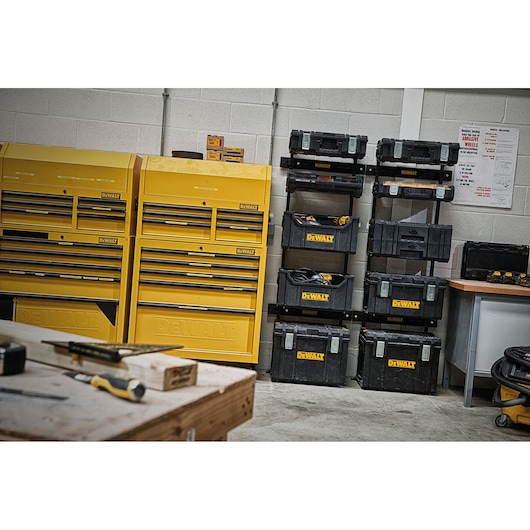 A workshop with several yellow DEWALT tool cabinets and a stack of black DEWALT toolboxes arranged neatly against a wall. There are various tools and equipment visible on a wooden workbench in the foreground.