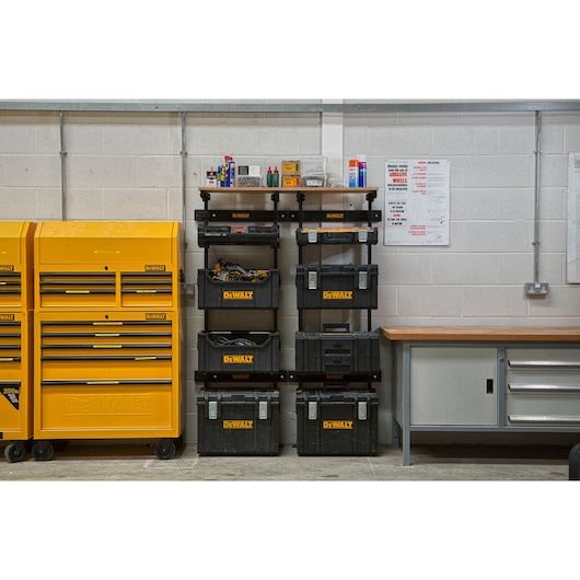 A workshop scene featuring DEWALT storage units, including yellow tool chests on the left and black modular toolboxes stacked on shelves in the center. The workspace is organized with tools, equipment, and supplies visible. A workbench with drawers is situated on the right, against a light gray wall.