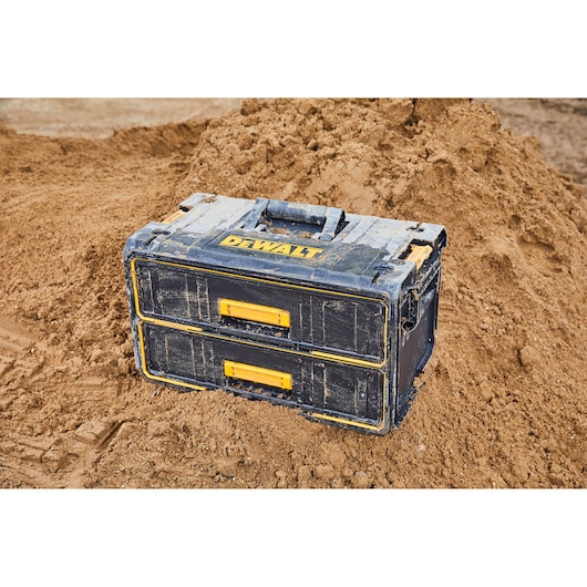 A DEWALT branded toolbox with two drawers and yellow handles, placed on a sandy construction site.