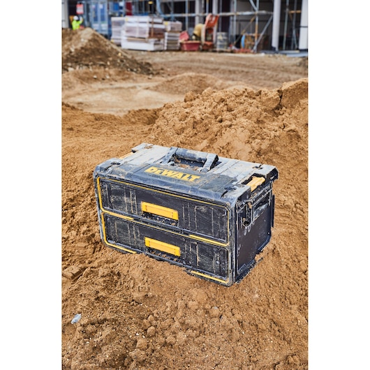 A rugged DEWALT tool storage box with two drawers, featuring yellow handles, placed on a construction site with dirt and building materials in the background.