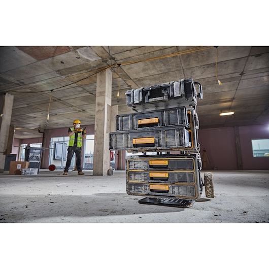 Stacked DEWALT toolboxes with yellow latches placed on a construction site floor, with a worker wearing a helmet and safety vest visible in the background.