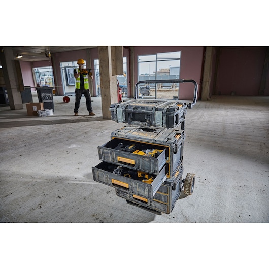 A DEWALT rolling tool storage cart with three open drawers, filled with power tools and accessories, is positioned on a construction site floor. A worker wearing a safety vest and helmet is visible in the background.