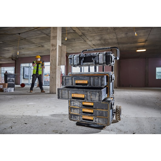 A stack of DEWALT toolboxes with yellow handles is shown on a rolling cart inside a construction site. A worker wearing safety gear is visible in the background.
