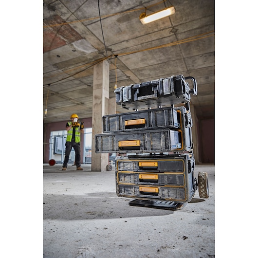 Stacked DEWALT tool storage boxes with yellow handles are shown on a wheeled cart at a construction site, with a worker in safety gear visible in the background.