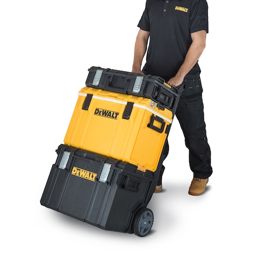A person is transporting a stack of DEWALT toolboxes on a wheeled cart. The toolboxes are black and yellow and have the DEWALT logo. The person is wearing a black DEWALT-branded shirt and black work pants.