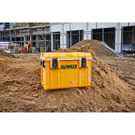 A yellow DEWALT tool storage box with black latches is placed on a pile of dirt at a construction site, with scaffolding and a building structure in the background.