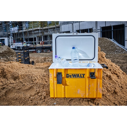 A yellow DEWALT cooler box with an open lid, containing plastic water bottles and ice, placed on a construction site with sand and building materials.