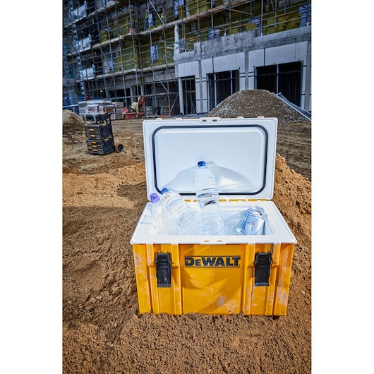 A yellow DEWALT cooler box with the lid open and several water bottles and cans inside, placed on a construction site with dirt piles and building scaffolding in the background.