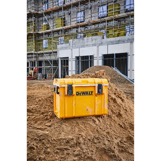 A yellow DEWALT storage box is placed on a mound of dirt at an outdoor construction site, with scaffolding and a building under construction visible in the background.