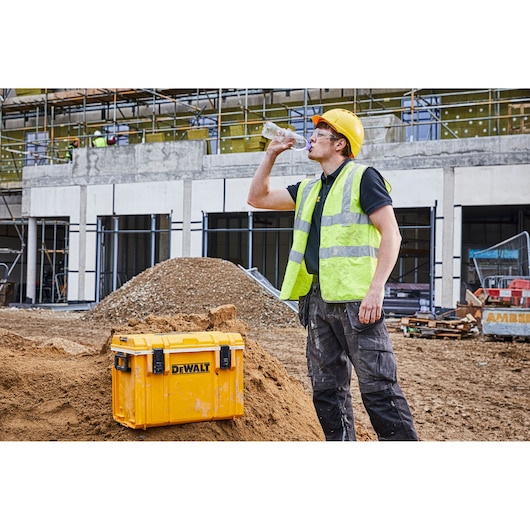 A person wearing a yellow hard hat and high-visibility vest is drinking water from a bottle at a construction site. In front of them is a yellow DEWALT toolbox placed on a pile of dirt. The background shows a building under construction with scaffolding and construction materials.