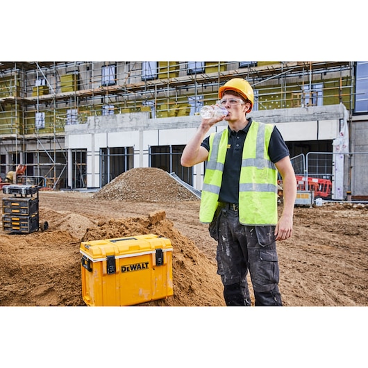 A person wearing a yellow safety vest and helmet stands at a construction site, drinking from a water bottle. In the foreground is a yellow DEWALT tool storage box (SKU: DWST1-81333) placed on a mound of dirt. Scaffolding and building materials are visible in the background.