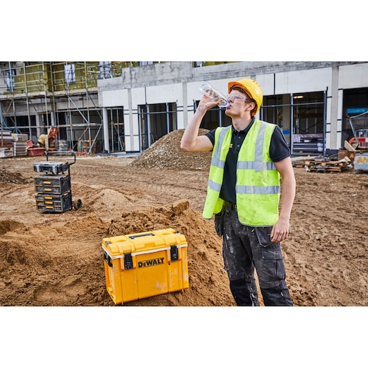 A person wearing a yellow hard hat and high-visibility vest is drinking from a water bottle at a construction site. In the foreground, there is a yellow DEWALT storage box placed on a mound of dirt, with additional DEWALT storage units and building materials visible in the background.