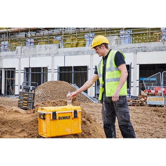A person wearing a yellow hard hat and reflective vest stands on a construction site next to a large yellow DEWALT toolbox (SKU: DWST1-81333) positioned on a mound of dirt. The person is placing a water bottle on the toolbox. Construction materials and another set of toolboxes are visible in the background.