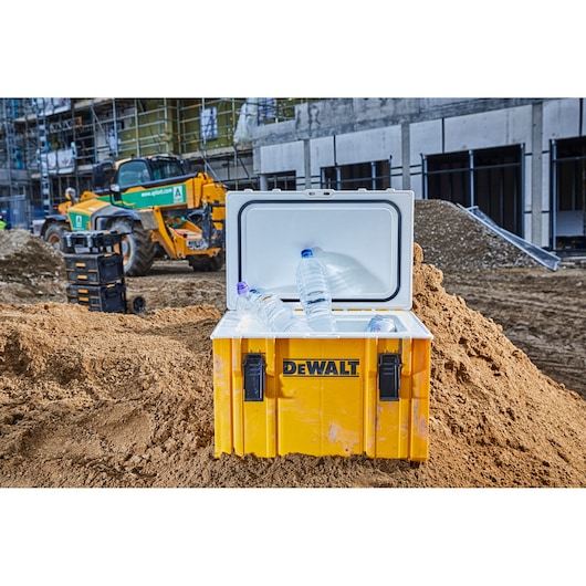A yellow DEWALT cooler box with an open lid, containing plastic water bottles, is placed on a dirt mound at a construction site. Construction vehicles and materials are visible in the background.