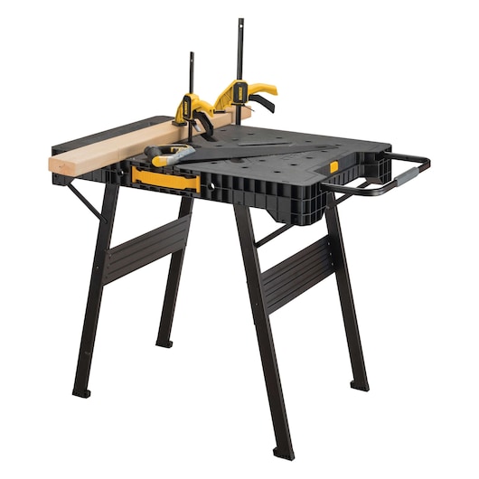 A DEWALT portable workbench with folding black legs, a robust black work surface, and yellow clamps holding a piece of wood in place. A saw and screwdriver are placed on top of the workbench.