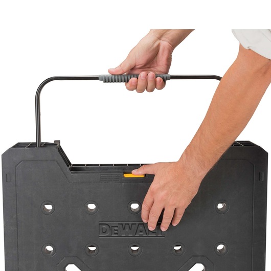 Close-up image showing hands holding and carrying a DEWALT-branded black plastic folding workbench with a handle.